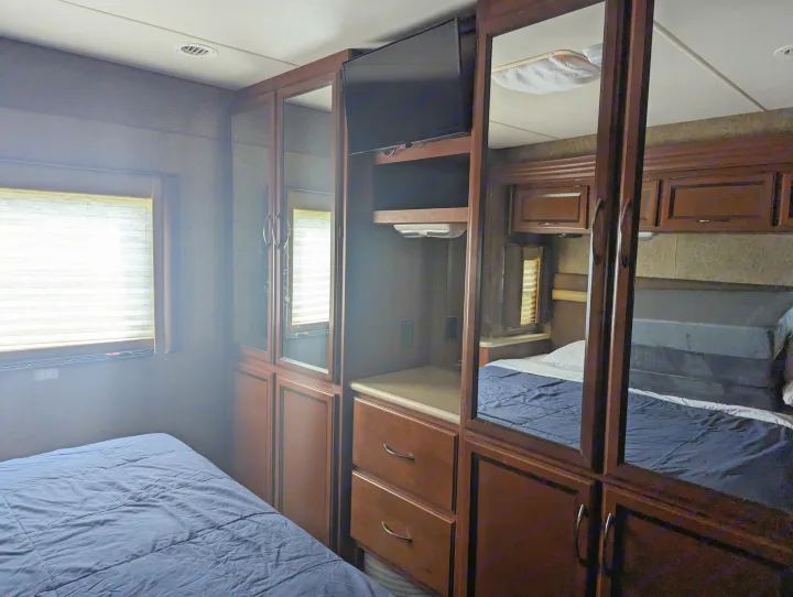Spacious RV rental bedroom with a comfy blue bed, window, and wood cabinetry featuring mirrors, TV, and storage.. Thor Other 2014