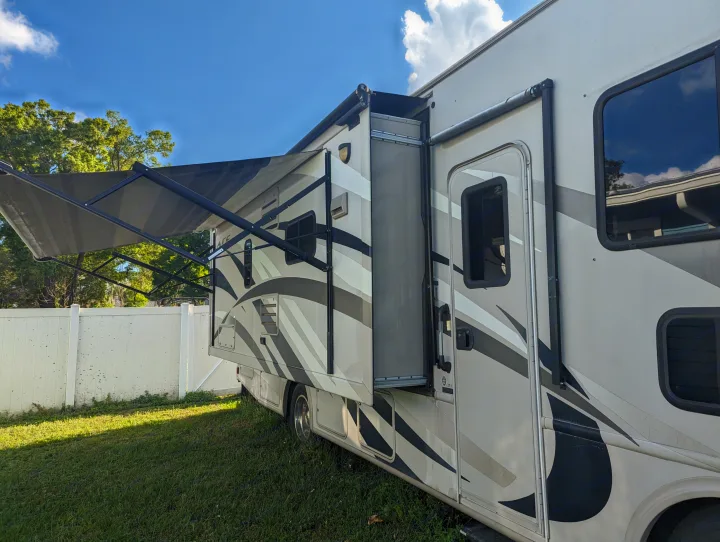 A light-colored RV rental with an extended awning and slide-out, parked on a sunny lawn next to a white fence.. Thor Other 2014