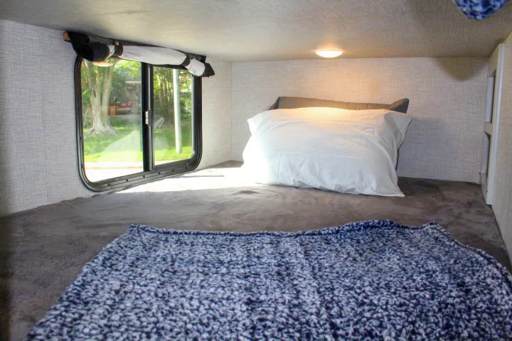 Inside an RV rental: a cozy sleeping bunk with a large window, a white pillow, and soft grey & blue blankets.. Keystone RV Bullet Crossfire 2021