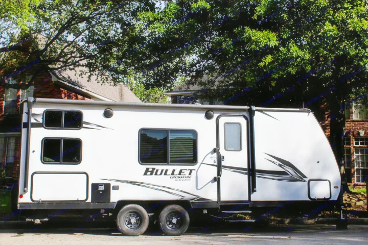 A large white Bullet Crossfire rv rental, a travel trailer, parked on a street with houses and green trees behind it.. Keystone RV Bullet Crossfire 2021