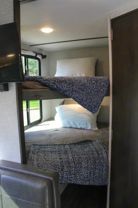 Inside an RV rental: cozy bunk beds with soft blankets & pillows, a TV, & window view. Perfect for travel comfort.. Keystone RV Bullet Crossfire 2021