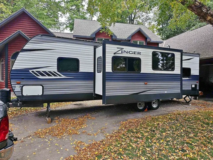 Zinger RV rental travel trailer parked in a driveway with autumn leaves beside a red house, slide-out extended.. Crossroads Zinger 2018