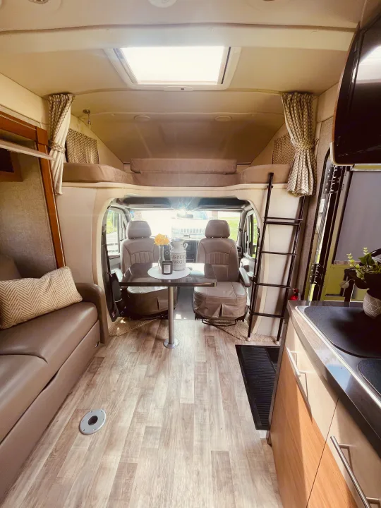 Inside a bright RV rental: features include kitchen, living space with sofa, dining table, front cockpit, and overhead bunk.. Winnebago View 2016