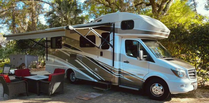 Modern Winnebago View RV, ideal for rv rental, parked on pavement with awning out, beside an outdoor patio set under lush trees.. Winnebago View 2016