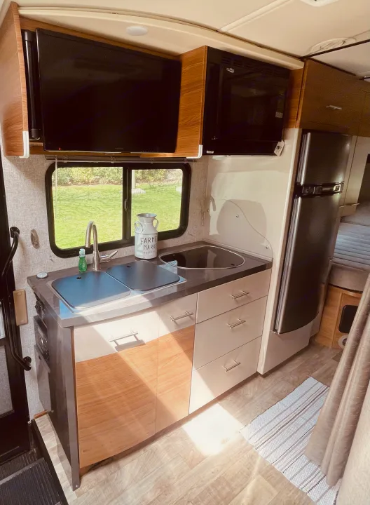 Clean and modern RV rental interior featuring a full kitchen with sink, microwave, fridge, and TV. Bright space with a view of nature.. Winnebago View 2016
