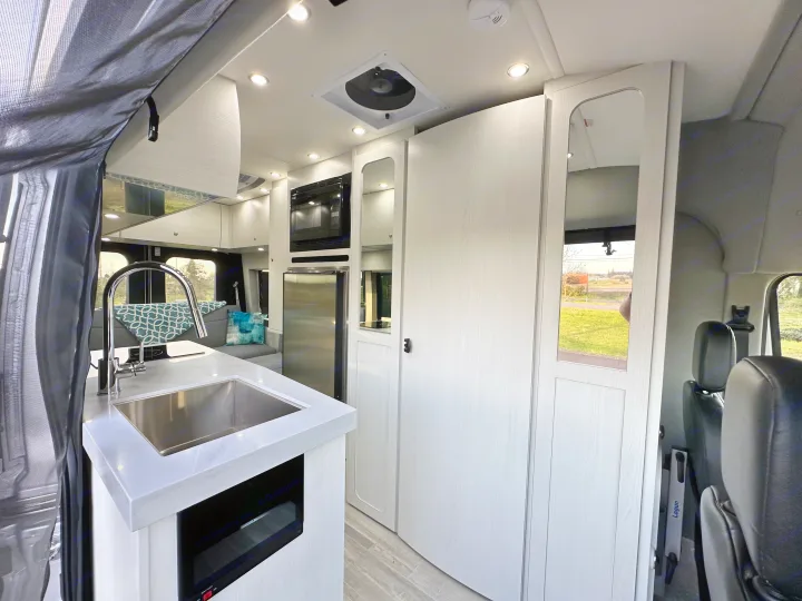 Modern RV rental interior: kitchen with sink, microwave, fridge, sleek white cabinets, and LED lighting. Bright and functional space.. Pleasure-Way Pleasure-Way Class B 2024