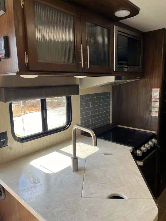 Modern RV rental kitchen with sleek countertops, sink, stove, microwave, and wood cabinets. Snowy outdoor view.. Forest River Cherokee Grey Wolf 2020