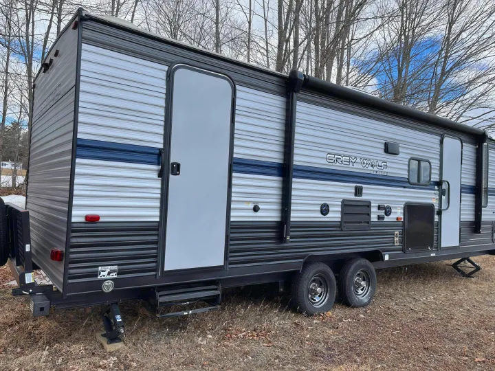 A grey travel trailer RV rental, featuring a blue stripe and awning, is parked on dry ground with bare trees behind it.. Forest River Cherokee Grey Wolf 2020