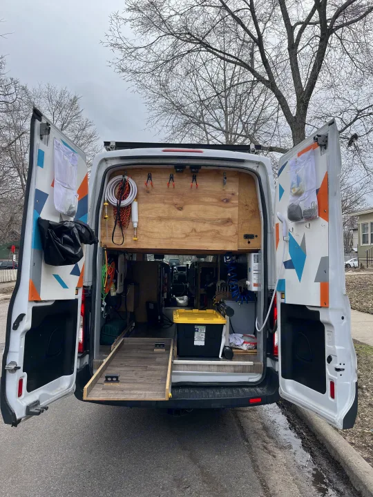 Open rear doors of an RV rental van reveal a custom-built interior with organized gear, an outdoor shower, and pull-out storage.. Ford Transit 2015
