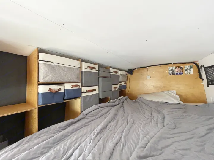 RV rental interior with a cozy bed, gray comforter, and efficient overhead storage in fabric bins on wooden shelves.. Ford Transit 2015