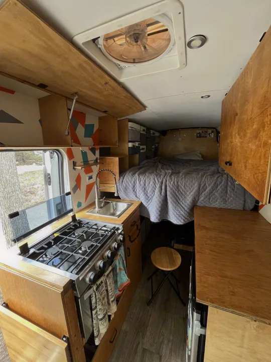 This RV rental offers a well-equipped camper van interior featuring a kitchen with stove/sink, bed, and overhead storage.. Ford Transit 2015