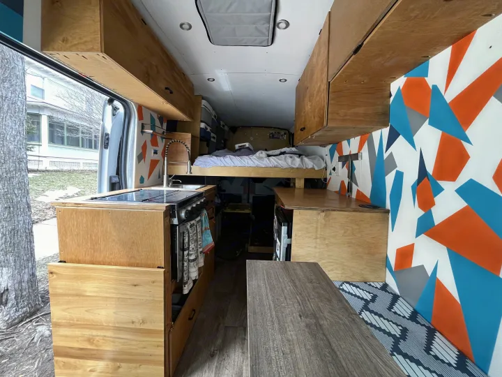 RV rental: View inside a modern camper van with a kitchen, raised bed, wood cabinetry, and vibrant geometric wall decor.. Ford Transit 2015