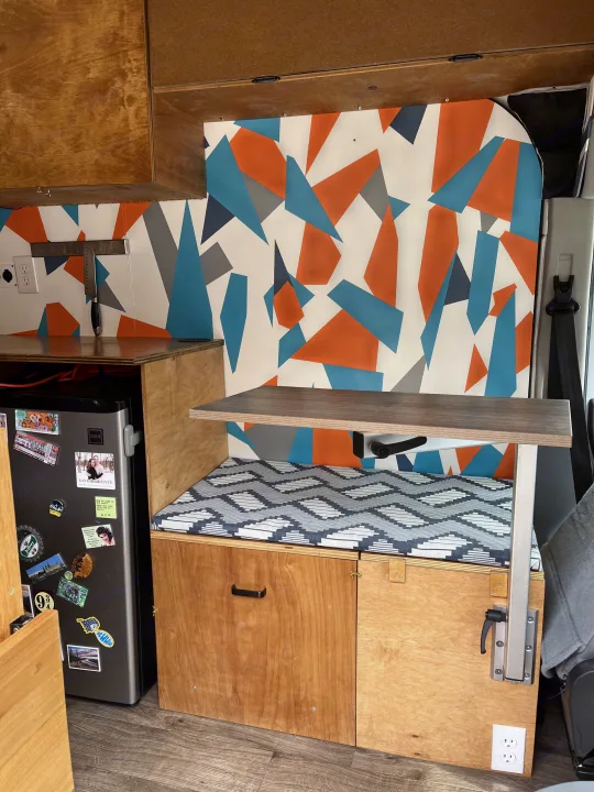 Stylish RV rental interior: wooden kitchenette with fridge, geometric accent wall, and a versatile fold-down table over seating.. Ford Transit 2015