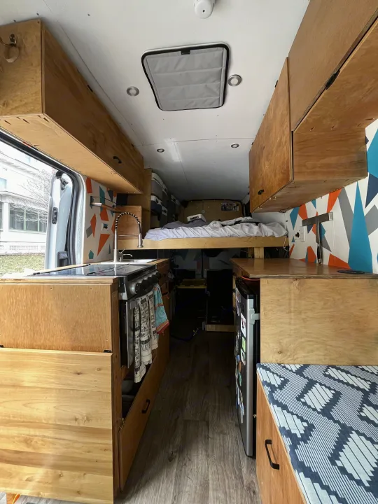 RV rental: Inside a modern camper van with a full kitchen, raised bed, fridge, and overhead cabinets.. Ford Transit 2015