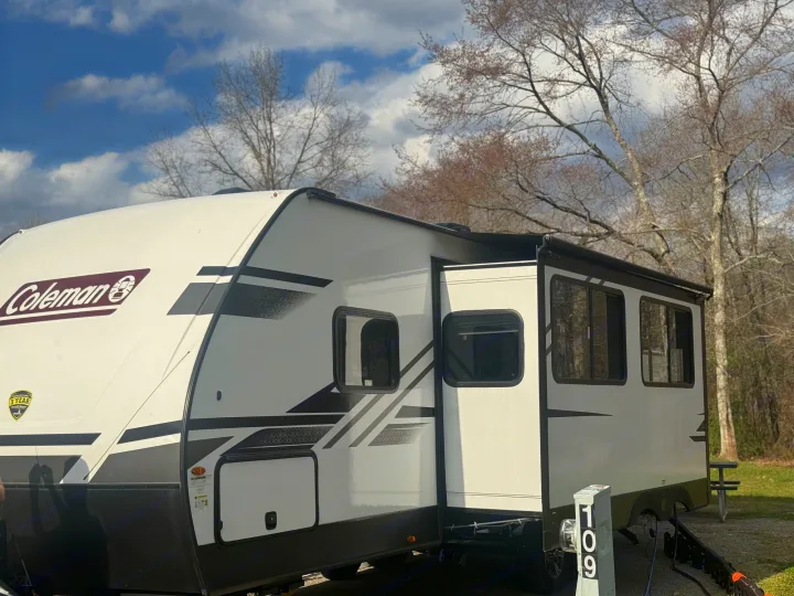 Modern white/black Coleman travel trailer at a sunny campsite, great for an rv rental. Blue sky, bare trees, and site 109 visible.. Dutchmen Coleman Light 2023