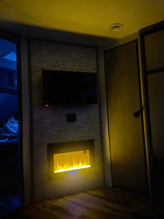 Inviting rv rental interior: TV over a brick-textured wall with glowing electric fireplace. Blue light from adjacent room.. Dutchmen Coleman Light 2023