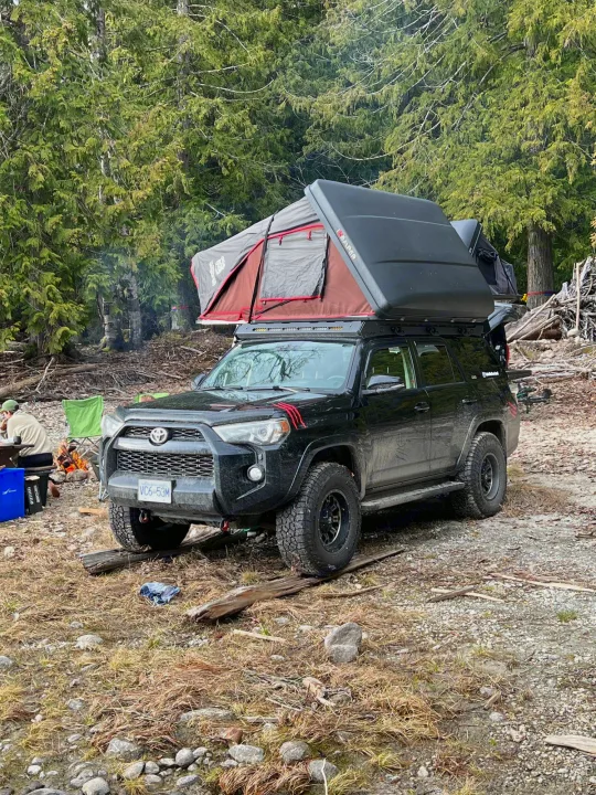 Black SUV with rooftop tent, a modern RV rental option for robust wilderness camping. Campfire glows nearby.. Toyota Other 2015