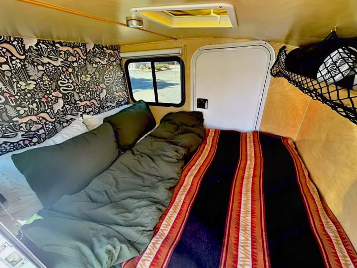 This cozy RV rental interior features a compact sleeping area with unique patterned wallpaper, a colorful blanket, and overhead storage.. Hiker Deluxe 2018