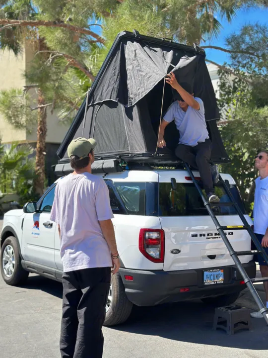 White Ford Bronco Sport with rooftop tent, a camping RV rental. Man exits tent via ladder as two others watch.. Ford Bronco Sport 2022