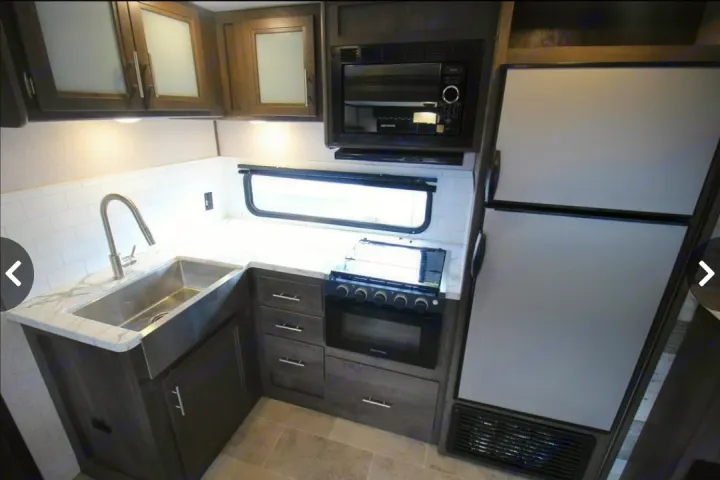 Modern RV rental kitchen with stainless sink, microwave, stove, oven, fridge, dark wood cabinets, and white subway tile backsplash.. Dutchmen Coleman Light 2020