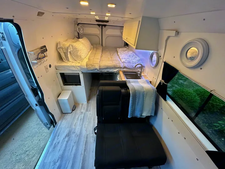 Spacious custom RV rental interior features a cozy bed, compact kitchen, and convertible seating with unique porthole windows.. RAM PROMASTER 2500 2023