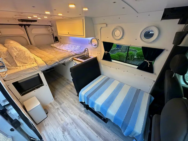 Inside an RV rental van: a cozy bed, compact kitchenette, and passenger seating area. Wood-look floors and ample lighting create a comfortable living space.. RAM PROMASTER 2500 2023