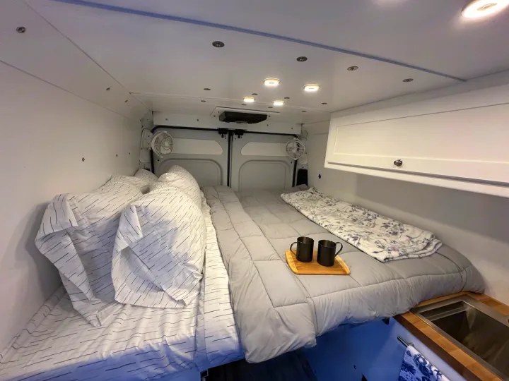 Experience comfort in this RV rental's sleeping area, featuring cozy bedding, overhead lights, fans, and a sink.. RAM PROMASTER 2500 2023
