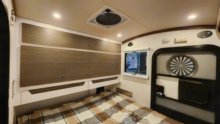 This RV rental interior features a cozy bedroom with a comfortable bed, wood-grain storage cabinets, window, ceiling fan, and door.. Bean Trailer Bean Trailer Trailer 2024