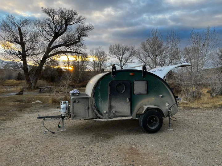This dusty, dark green teardrop camper is set up at a scenic campsite during sunset, perfect for your next RV rental adventure.. Bean Trailer Bean Trailer Trailer 2024