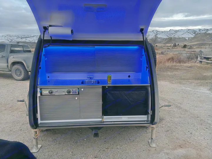 Teardrop RV rental: Illuminated outdoor kitchen with sink, stove, and storage. Night camp scene with snowy mountains.. Bean Trailer Bean Trailer Trailer 2024