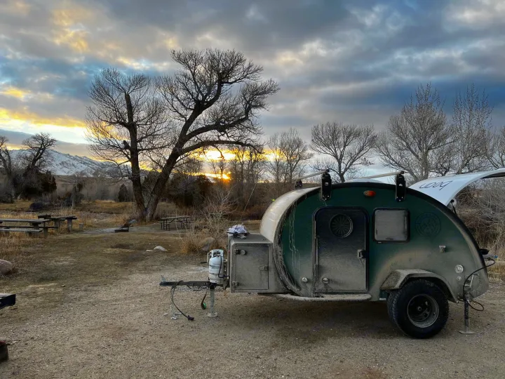 Dusty green teardrop RV rental at a campsite with bare trees and a vibrant sunset behind mountains. Perfect for off-grid camping!. Bean Trailer Bean Trailer Trailer 2024