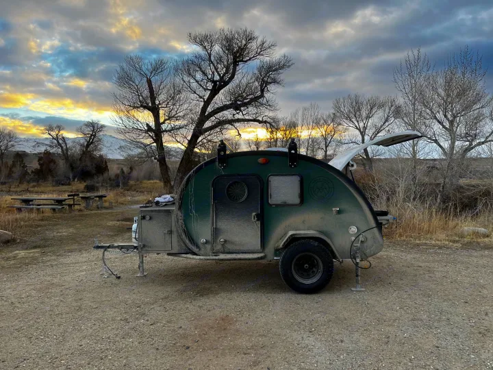 This rugged green teardrop RV rental is set against a vibrant sunset sky and mountains, ready for camping adventures.. Bean Trailer Bean Trailer Trailer 2024