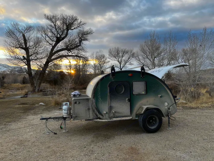 A dusty, green teardrop RV rental is parked in a rustic campground at sunset, surrounded by bare trees and mountains.. Bean Trailer Bean Trailer Trailer 2024
