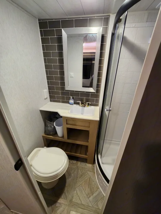 RV rental bathroom: A compact space with a toilet, vanity with sink, and curved glass shower. Features modern grey tiling & wood accents.. Forest River Wildwood 2023