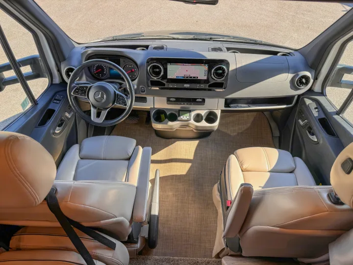 Detailed view of a modern RV rental's driver & passenger cockpit, showing dashboard, steering wheel, infotainment, & seating.. Thor Motor Coach Delano 2022