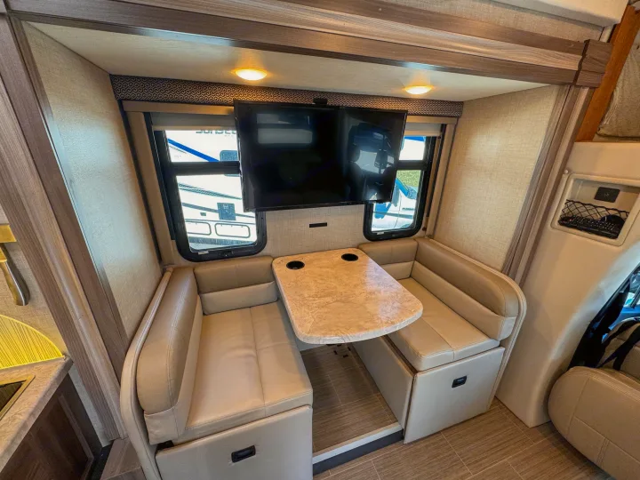 Inside an RV rental: a cozy dinette with tan upholstered seating, a table with cupholders, and a mounted TV.. Thor Motor Coach Delano 2022
