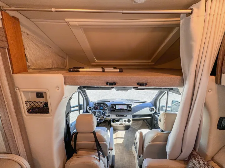 RV rental interior looking forward from the living area to the driver's cab, showing an overhead bed and dashboard.. Thor Motor Coach Delano 2022