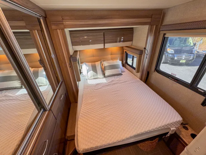 Inside a modern RV rental: a cozy bedroom with a quilted bed, overhead cabinets, and a window view to the outdoors.. Thor Motor Coach Delano 2022