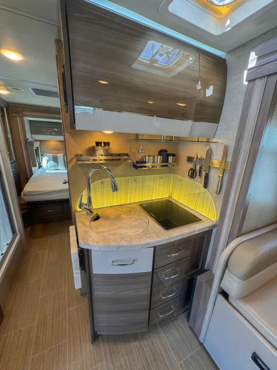 Modern RV rental interior: compact kitchen with sink, cooktop, and view to a cozy bedroom. Ideal for travel comfort.. Thor Motor Coach Delano 2022