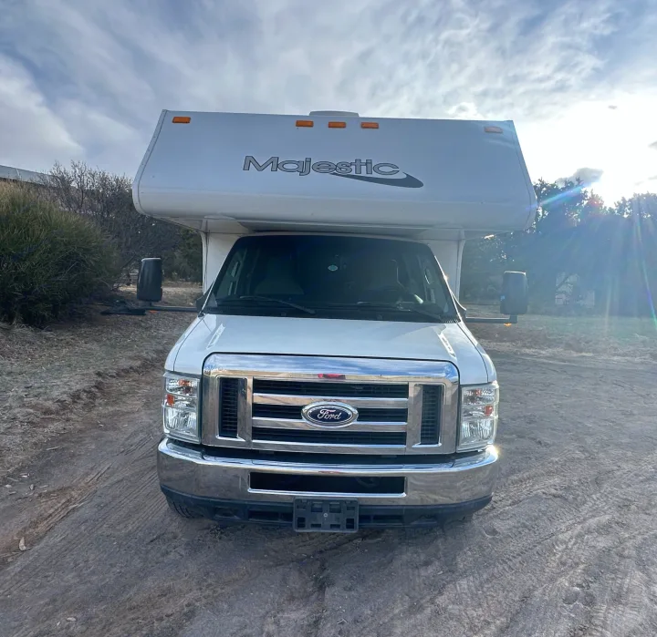 Front view of a white Majestic Class C Ford RV rental, parked outdoors on dirt under a cloudy sky with sun glare.. Thor Majestic 2016