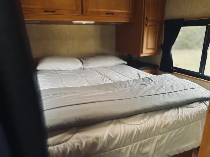 Cozy interior of an RV rental bedroom with a neatly made bed, white and gray bedding, wooden cabinets, and a window.. Thor Majestic 2016