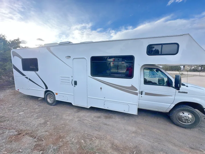 Full view of a white Class C RV rental with graphic stripes parked on an unpaved surface under cloudy skies.. Thor Majestic 2016