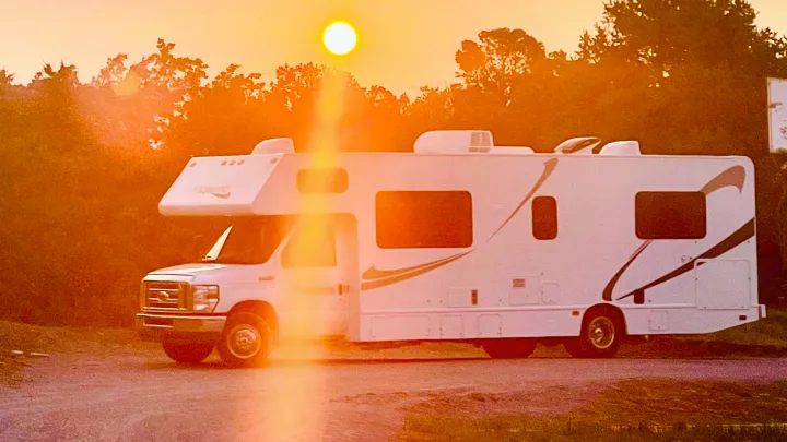 A white Class C rv rental glows under a golden sunset, parked on a dirt road with silhouetted trees and sun flare.. Thor Majestic 2016