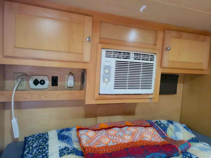 RV rental bedroom: a cozy bed below wooden cabinets, AC, and a shelf with various electrical outlets.. Little Guy Silver Shadow 5x10 2015