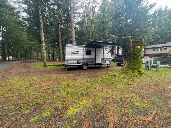 A silver Coleman travel trailer is parked at a mossy campground site #107 amidst tall trees, perfect for an RV rental.. Coleman 17B 2024
