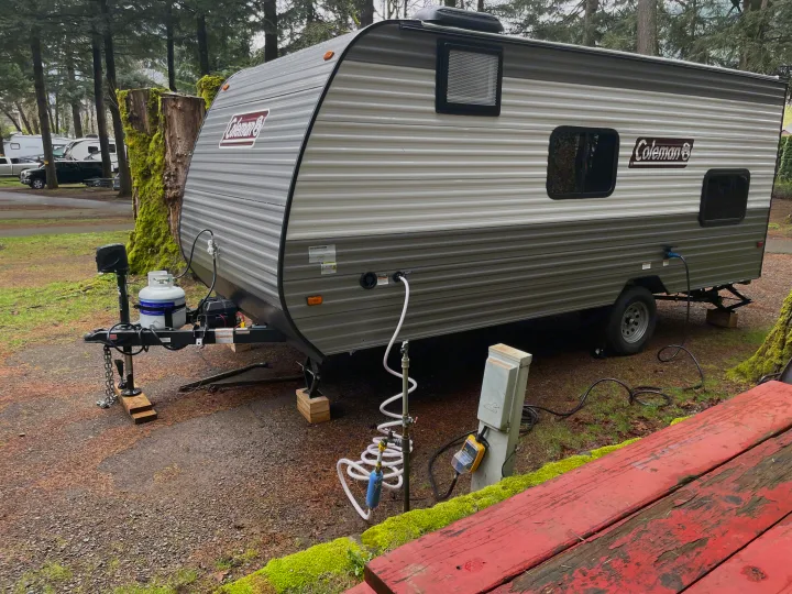 Grey Coleman RV rental set up at a wooded campsite, showing propane, water, and power hookups, with a red table.. Coleman 17B 2024