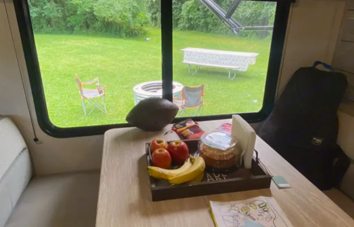 Relaxing inside an RV rental: Dinette with snacks and fruit overlooking a campsite with a fire pit, picnic table, and chairs.. Prime Time Avenger by Forest River 26bk 2023