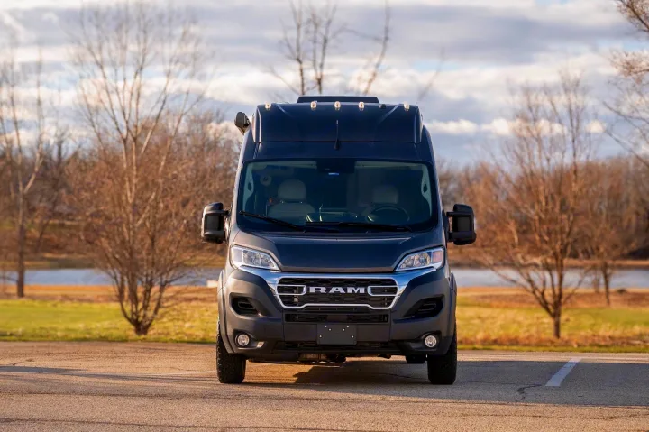 This front view shows a dark grey Ram ProMaster RV rental van parked in a natural setting with trees and water.. Thor Sequence 2024