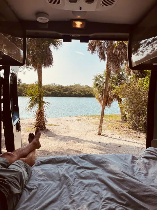 Ultimate relaxation in an RV rental: feet up on the bed, enjoying a scenic view of tranquil water and palm trees from inside the van.. Thor Sequence 2024