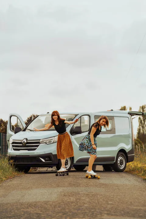 Two women smile on skateboards by their light blue RV rental camper van, ready for a road trip adventure.. Renault Trafic 2025